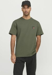 Camiseta de algodón verde oliva con mangas cortas, cuello redondo y un pequeño logo gráfico negro/rojo en el lado izquierdo del pecho. Modelo lleva una gorra negra.