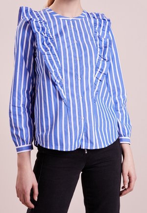 Femme portant un chemisier à rayures verticales bleues et blanches avec des volants à l'avant et un pantalon noir, debout devant un fond clair.