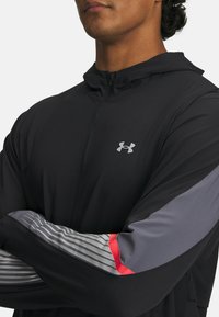 Schwarze Zipjacke mit einem grauen und gestreiften Ärmel-Design, das einen roten Akzent aufweist. Das Material wirkt leicht und glatt.