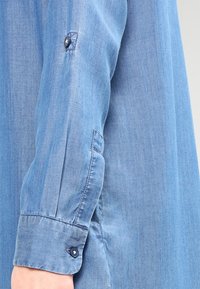 Ljusblå denimskjorta med ärm som har knäppning och manschettdetaljer, som visar en jämn textur och subtila sömmar.