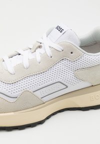 Sneaker in rete bianco e suede beige con accenti grigi. Presenta lacci rotondi e un logo sulla linguetta e sul lato. Suola testurizzata per maggiore aderenza.