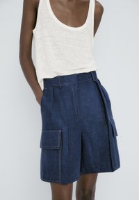 Personne portant un short cargo en denim bleu foncé et un haut blanc sans manches ample, une main dans la poche.