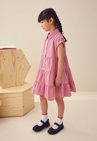 Vestito a quadretti rossi con maniche corte, balze a più strati e bottoni frontali. Indossato con scarpe scolastiche nere in vernice e calzini bianchi con bordino rosso.