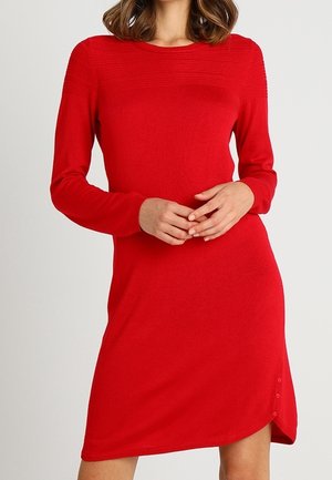 Femme portant une robe rouge ajustée à manches longues, avec un col rond et une petite fente avec des boutons sur le côté inférieur.