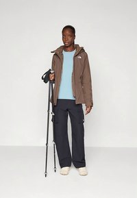 Barna vízálló dzseki kapucnival, világoskék ing, sötét cargo nadrágok és túrabotok. Puha bézs lábbeli. Minimalista kialakítás.