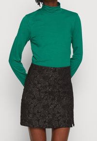 Grön långärmad polokrage topp kombinerad med en svart minikjol med ett texturerat blommönster. Ensemblet är figursydd och modernt.