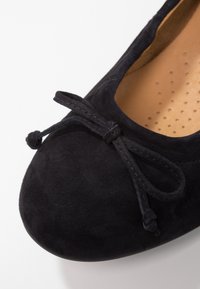 Ballerines en daim noir avec un bout rond, un petit nœud sur le devant, et un intérieur lisse avec des perforations.