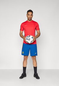 Camiseta de manga curta vermelha da Nike, calções azuis com emblema amarelo, segurando uma bola de futebol estampada, meias pretas e botas de futebol pretas.