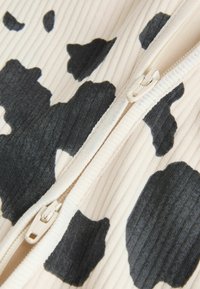 Schwarz-cremefarbenes, geripptes Stoffmaterial mit Kuhfleckenmuster. In der Mitte befindet sich ein cremefarbener Reißverschluss. Die strukturierte Oberfläche sorgt für visuelles Interesse.