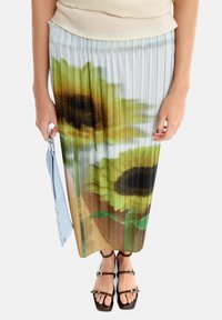 Maxi spódnica z plisowaniem, ozdobiona printem słonecznika, uszyta z miękkiego materiału w odcieniach zieleni i brązu. Połączona z czarnymi sandałami na paski i niebieską torbą.