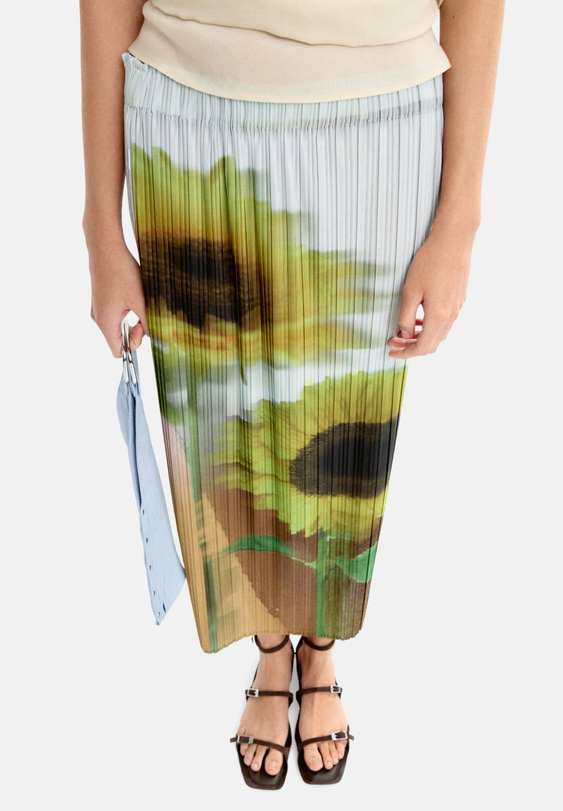 Maxi spódnica z plisowaniem, ozdobiona printem słonecznika, uszyta z miękkiego materiału w odcieniach zieleni i brązu. Połączona z czarnymi sandałami na paski i niebieską torbą.