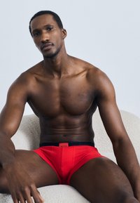 Sarkanas pieguļošas peldbikses ar melnu jostasvietu, demonstrētas uz muskuļota ķermeņa modeļa, kas sēž uz gaišas krāsas teksturēta krēsla.