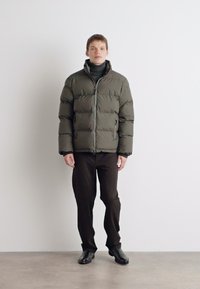 Jachetă tip puffer de culoare verde măsliniu, cu textură matlasată și fermoar frontal. Asortată cu pantaloni închis la culoare și pantofi negri, pe un fundal neutru.