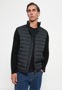 Young man with slicked-back hair wearing a black puffer vest over a black long-sleeve shirt and blue jeans, standing against a plain white background.