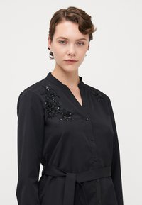 Abito camicia nero con colletto, caratterizzato da ricami floreali in perline sulle spalle, chiusura con bottoni e una cintura di tessuto in vita. Texture morbida.