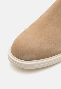 Botín de ante beige hasta el tobillo con punta redonda y suela de goma blanca, que presenta una textura suave y elementos de diseño minimalistas.