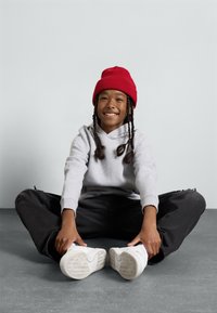 Felpa con cappuccio grigia, pantaloni neri oversize e sneakers bianche. Un bambino è seduto con le gambe incrociate, indossando un berretto rosso e sorridendo.