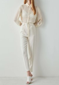 Blouse légère de couleur crème, plissée, avec col et boutons, associée à un pantalon taille haute, ajusté, et des mules à talons ouvertes.