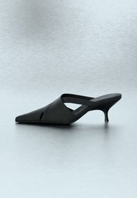 Sort mule med spids tå, lav kittenhæl og bred rem, der krydser over foden, på en neutral baggrund.