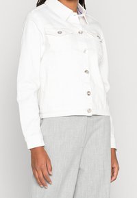 Biała kurtka dżinsowa z kołnierzykiem, z zapięciem na guziki, dwoma kieszeniami na piersi i srebrnymi detalami. Nosi się z jasnoszarymi spodniami. Gładka tekstura.