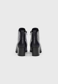 Une paire de bottines en cuir noir avec des talons blocs et des détails de fermeture éclair à l'arrière, montrées de l'arrière sur un fond blanc.