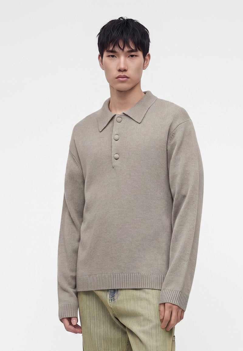 Jeune homme aux cheveux noirs courts portant un pull en maille beige avec col et trois boutons, debout devant un fond blanc uni.