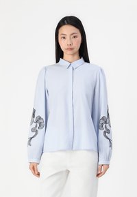 Camisa de manga longa azul clara com colarinho abotoado e bordado floral preto nas mangas. Tecido suave com uma textura lisa.