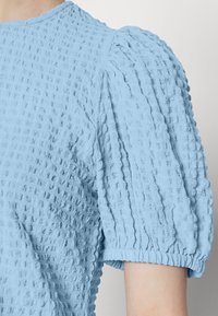 Top em azul claro com textura, mangas bufantes, padrão de xadrez, punhos elásticos e decote redondo. A textura do tecido é visível.