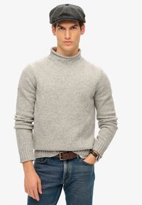 Grauer Strickpullover mit Rollkragen und gerippten Bündchen, kombiniert mit blauen Jeans. Eine dunkle Schirmmütze und eine Uhr runden das Outfit ab.