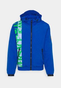 Blå puffjacka med huva, med en grön och blå logotyptryck på sidan, dragkedja fram och elastiska ärmslut. Slät textur.