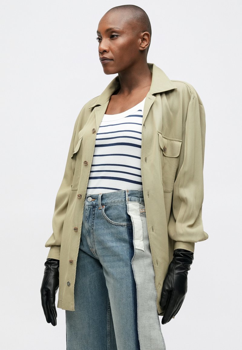 Olivgrön skjorta med knäppning och två bröstfickor, kombinerad med en randig linne och vida, lapptäckta denimjeans. Svart läderhandskar.