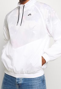 Windbreaker bianco Nike SB con collo mezza zip, cordini neri, polsini elastici e finitura strutturata, indossato sopra jeans in denim.