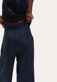 Pantalons en lin navy à jambe large, avec poches arrière et passants de ceinture, présentés de l'arrière, avec des mains ajustant la taille.