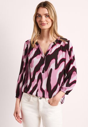 Femme aux cheveux blonds portant un chemisier à manches longues avec des motifs abstraits roses, noirs et blancs, associé à un pantalon blanc, main dans la poche.