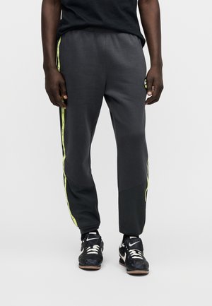 Mann trägt schwarze Sporthose mit neongelben Seitenstreifen und schwarze Nike-Sneaker mit weißen Akzenten, steht vor einfarbigem Hintergrund.