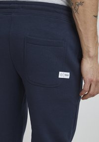 Marineblaue Sweatpants mit einer Gesäßtasche und einem weißen Etikett, das mit dem Markenlogo geprägt ist. Der Stoff wirkt weich und glatt.