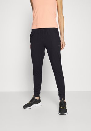 Kvinde iført sorte løbebukser, sort-hvide sneakers og en ærmeløs ferskenfarvet top, der står mod en ensfarvet lys baggrund.