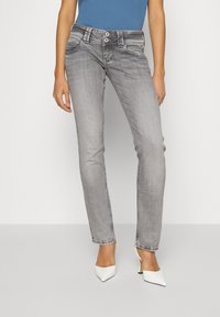 Jeans de mezclilla gris con un ajuste slim, costuras visibles, dos bolsillos frontales, cierre de botón y textura desgastada. Combinados con tacones blancos.