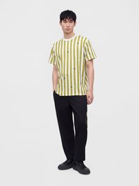 Un jeune homme se tient debout, les mains partiellement dans les poches, portant un t-shirt blanc avec des bandes verticales jaunes en forme de ruban à mesurer et un pantalon et des chaussures noirs.