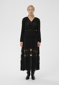 Čierne plisované šaty s dlhými rukávmi, výstrihom do V a vrstvenými priehľadnými časťami. Tkanina má prúdiacu textúru. Nosené s čiernymi čižmami po kolená.