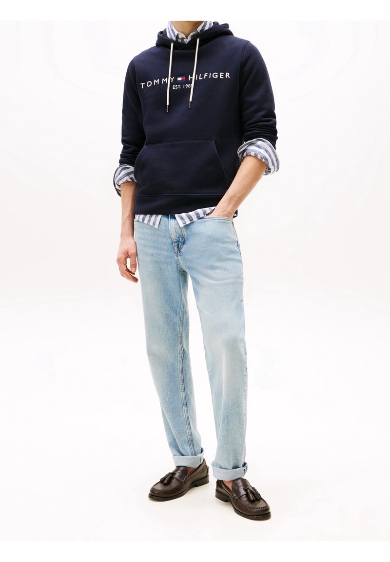 Felpa con cappuccio blu marina con logo bianco, jeans azzurri chiari e mocassini scuri. La camicia sotto presenta un motivo a righe blu e bianche.