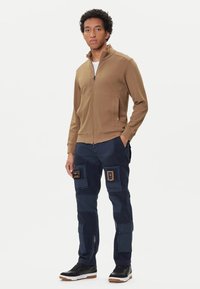 Giacca marrone con zip e pantaloni cargo blu navy con molte tasche e toppe. Sneakers nere con accenti bianchi e marroni. Tessuto morbido.