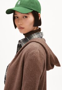 Brauner gestrickter Cardigan mit Kapuze, über einem karierten Hemd getragen. Die Person trägt eine grüne Mütze mit einem gestickten Logo.