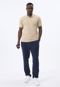 Beige Poloshirt mit kurzen Ärmeln und einer Knopfleiste mit drei Knöpfen, kombiniert mit marineblauen Hosen und weißen Sneakern. Glatte Baumwollstruktur.