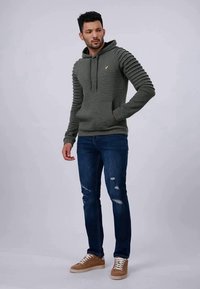 Felpa verde texturizzata con maniche a coste, tasca frontale e coulisse. Indossata con jeans blu strappati e sneakers beige.