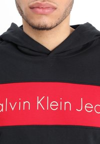 Sudadera negra con una banda roja que presenta el texto blanco "Calvin Klein Jeans". Tela suave con capucha y un ajuste relajado, adecuada para el uso diario.