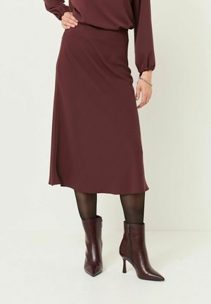 Vrouw die een bordeaux midi-rok draagt, bijpassende top, zwarte doorzichtige panty's en bordeaux puntige enkellaarzen met hak, staand met de hand op de heup.