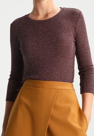 Femme portant un haut ajusté à manches longues de couleur bordeaux foncé avec une texture scintillante et une jupe taille haute jaune moutarde avec des poches.