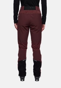 Person, der burgunderfarbene und schwarze Outdoor-Hosen trägt, steht von hinten mit schwarzen Schuhen und einem schwarzen Langarmoberteil.