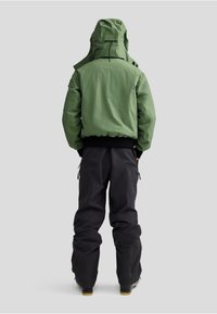 Veste imperméable verte avec capuche, ceinture élastique et pantalons de ski noirs. La veste présente une texture lisse et des cordons de serrage réglables.